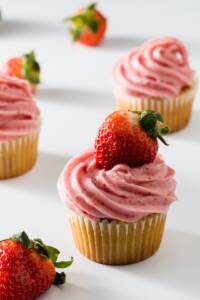 A group of cupcakes on white topped with strawberry cream cheese frosting topped with a fresh strawberry
