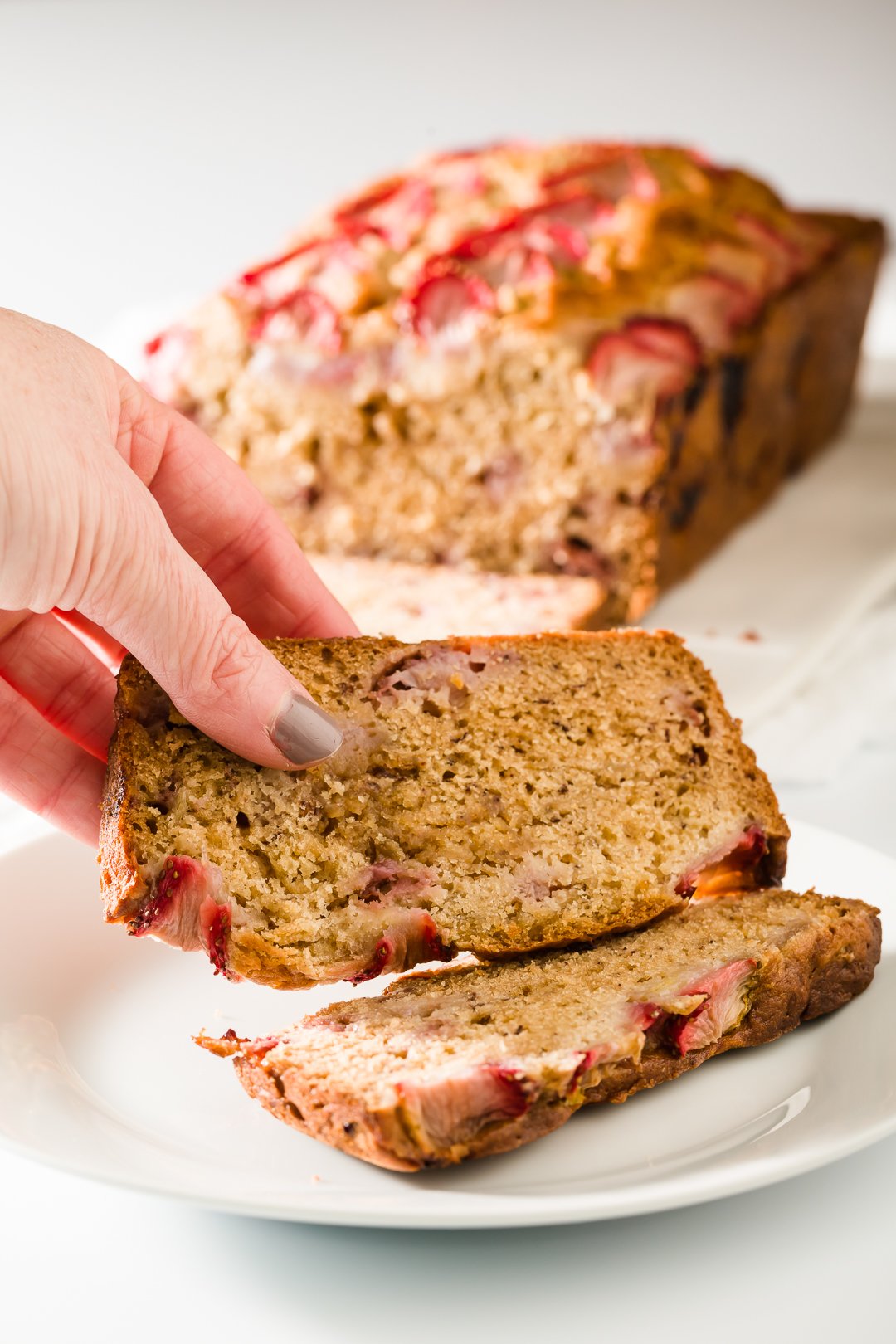A hand holding a slice of strawberry banana bread on a white plate with a loaf of strawberry banana bread in the background