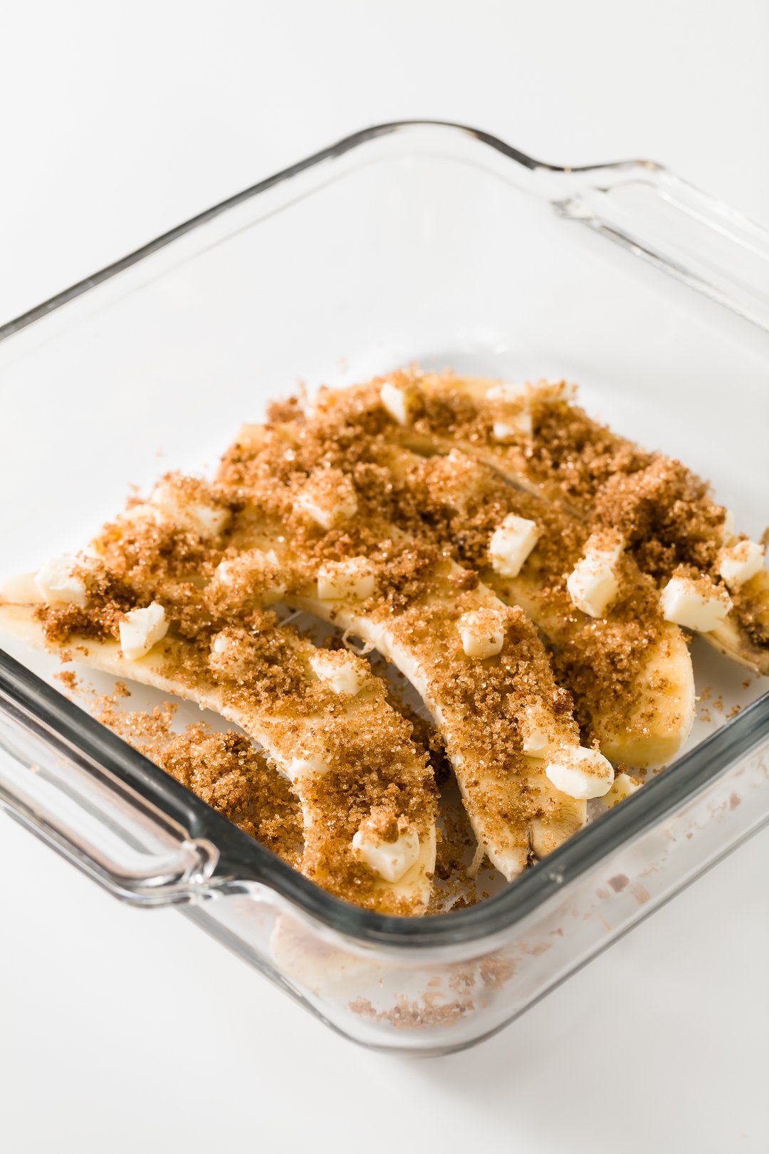 Bananas sliced longways in a glass baking dish covered in butter and brown sugar