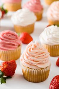 cupcakes frosted with three types of strawberry frosting on a white background with some scattered strawberries