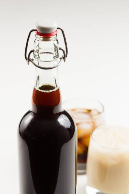 una botella de Kahlua casera en primer plano con dos cócteles hechos al fondo
