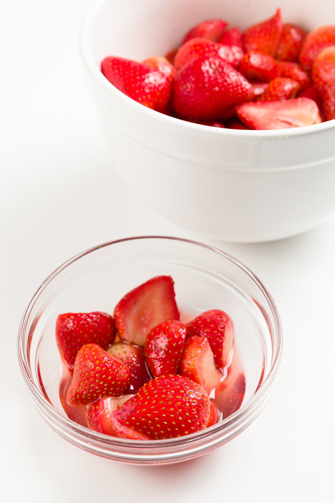 A small bowl of glistening macerated strawberries with a larger bowl full of strawberries in the background