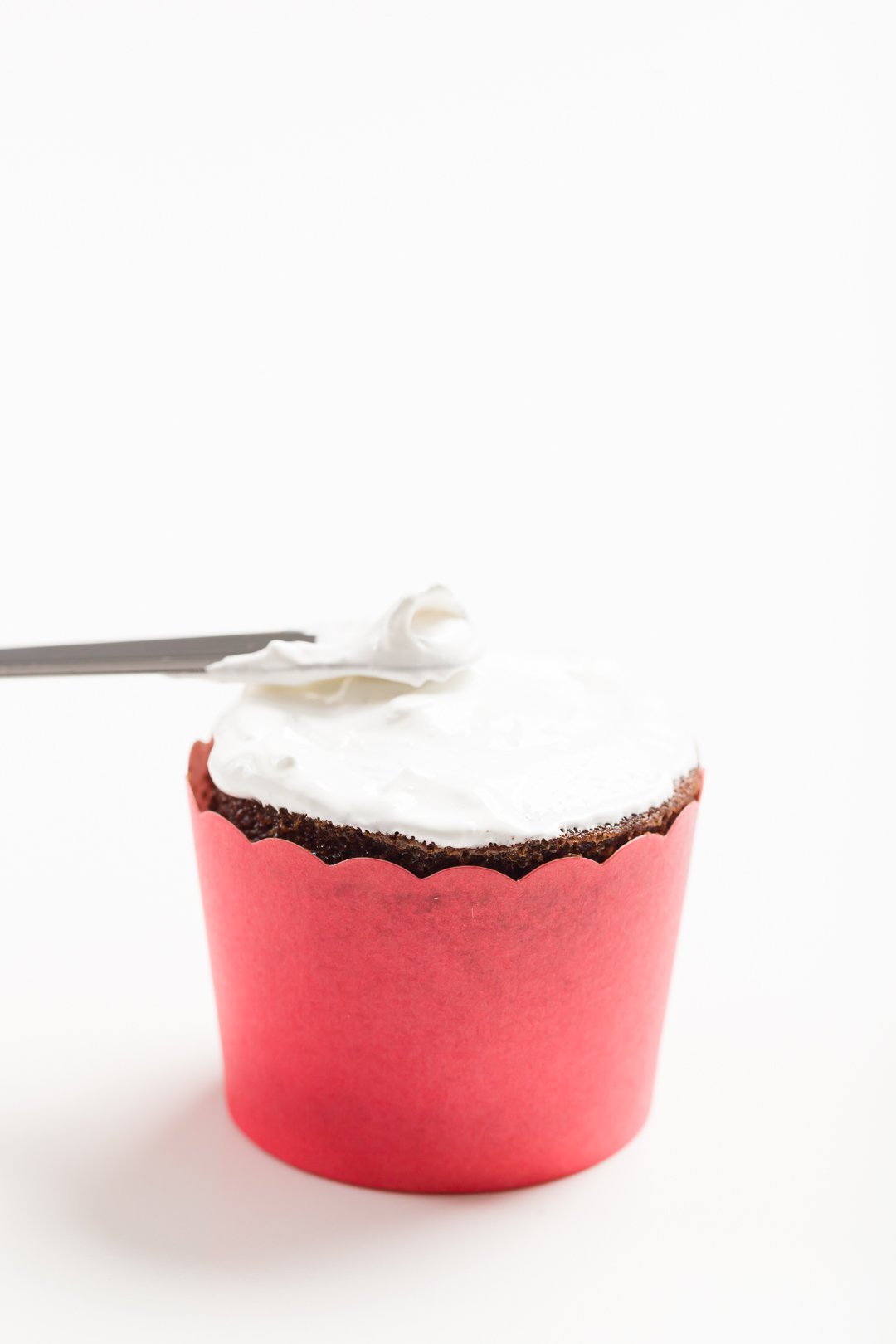 head-on view of spreading a thin layer of marshmallow frosting on top of a chocolate cupcake in a red baking cup