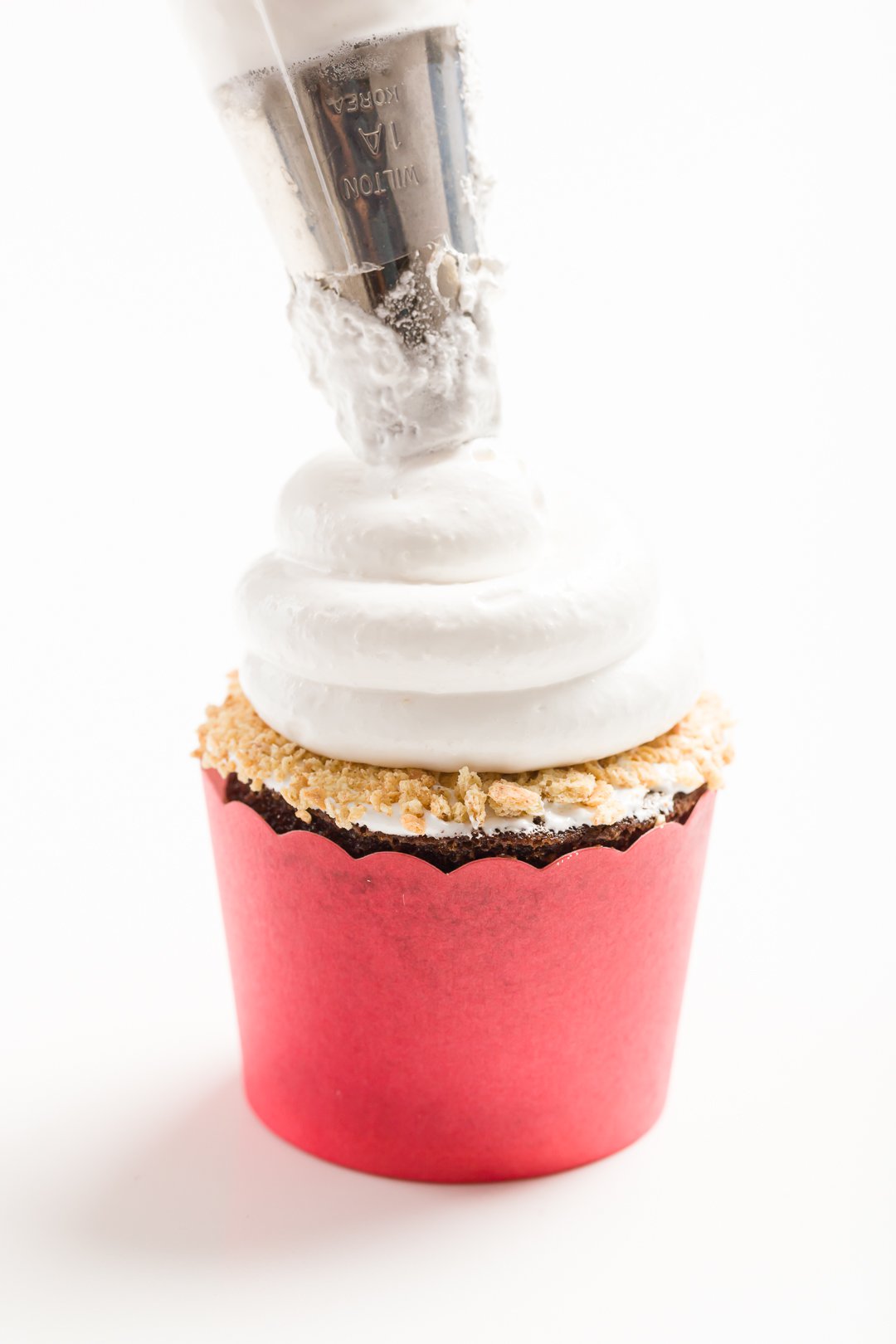 head-on view of piping marshmallow frosting onto a a s'mores cupcake in a red baking cup