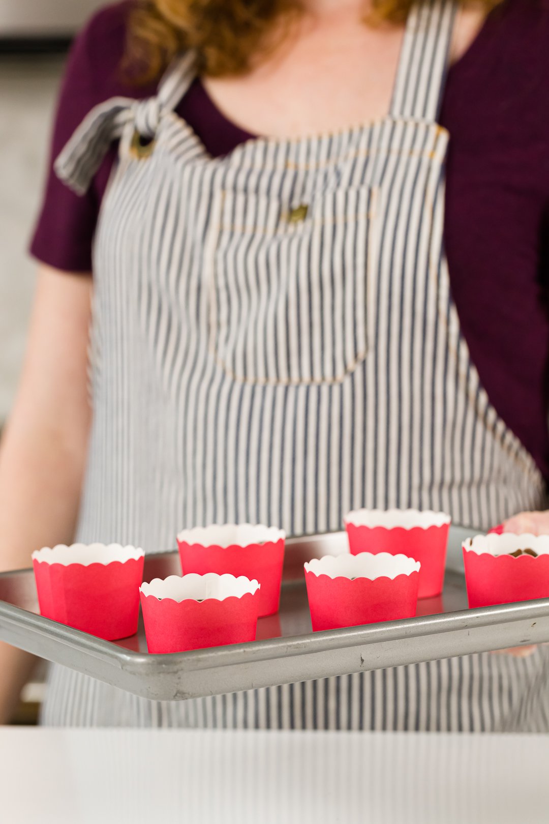 Blurred out person in a striped apron holding a cooking sheet with red baking cups on it