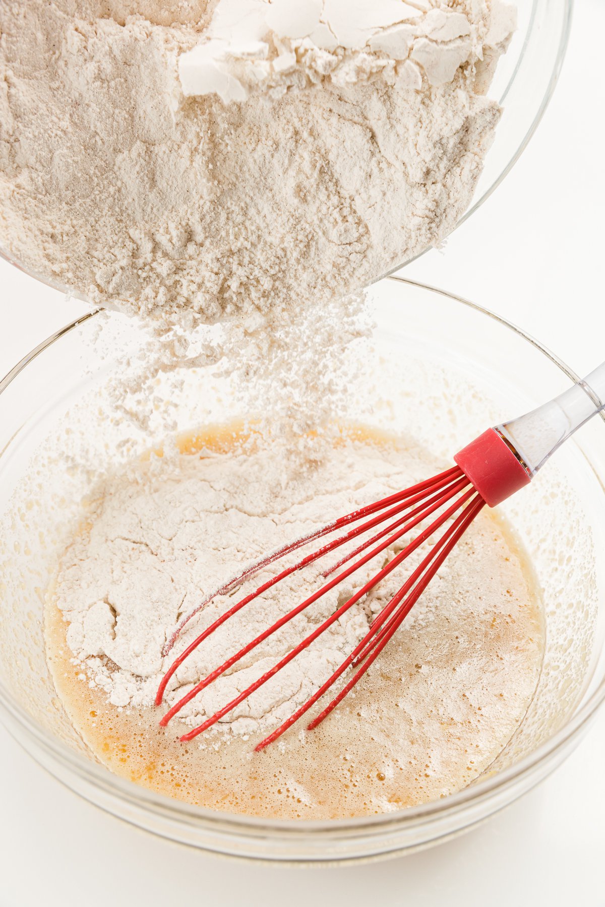 Flour pouring into a bowl of wet ingredients with a red whisks