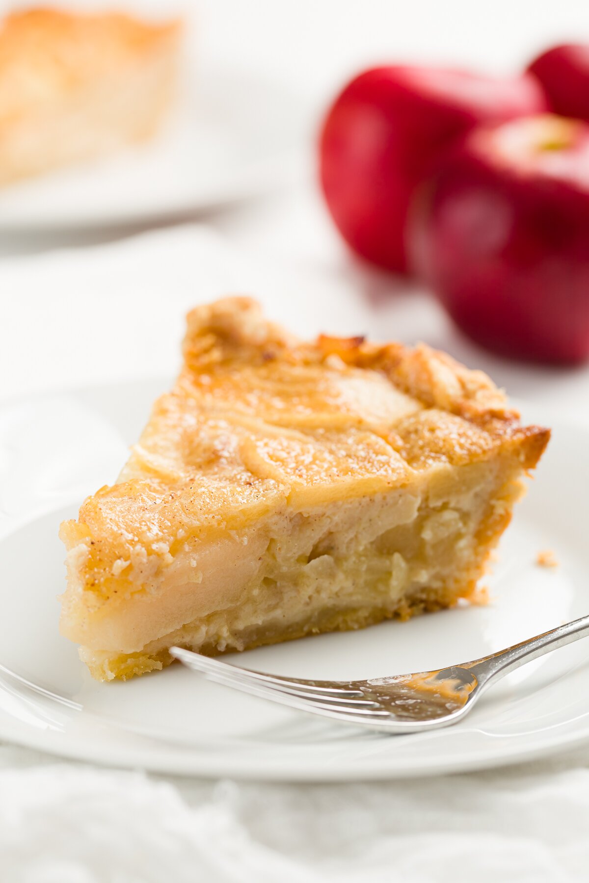 slice of apple custard pie on a white plate with a fork and apples in the background