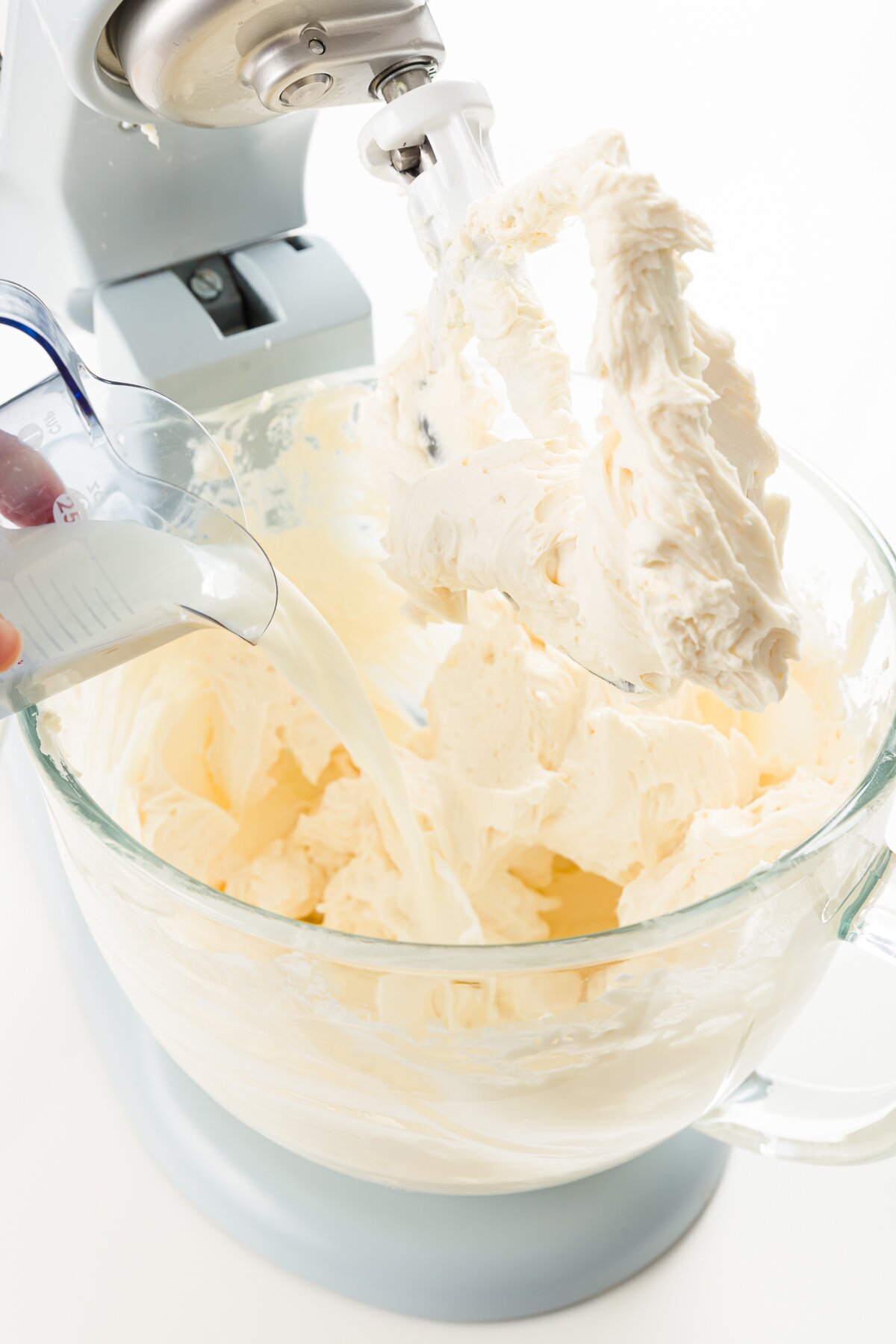 Adding milk to mixer bowl of a stand mixer with lifted paddle full of cream cheese and sugar mixture