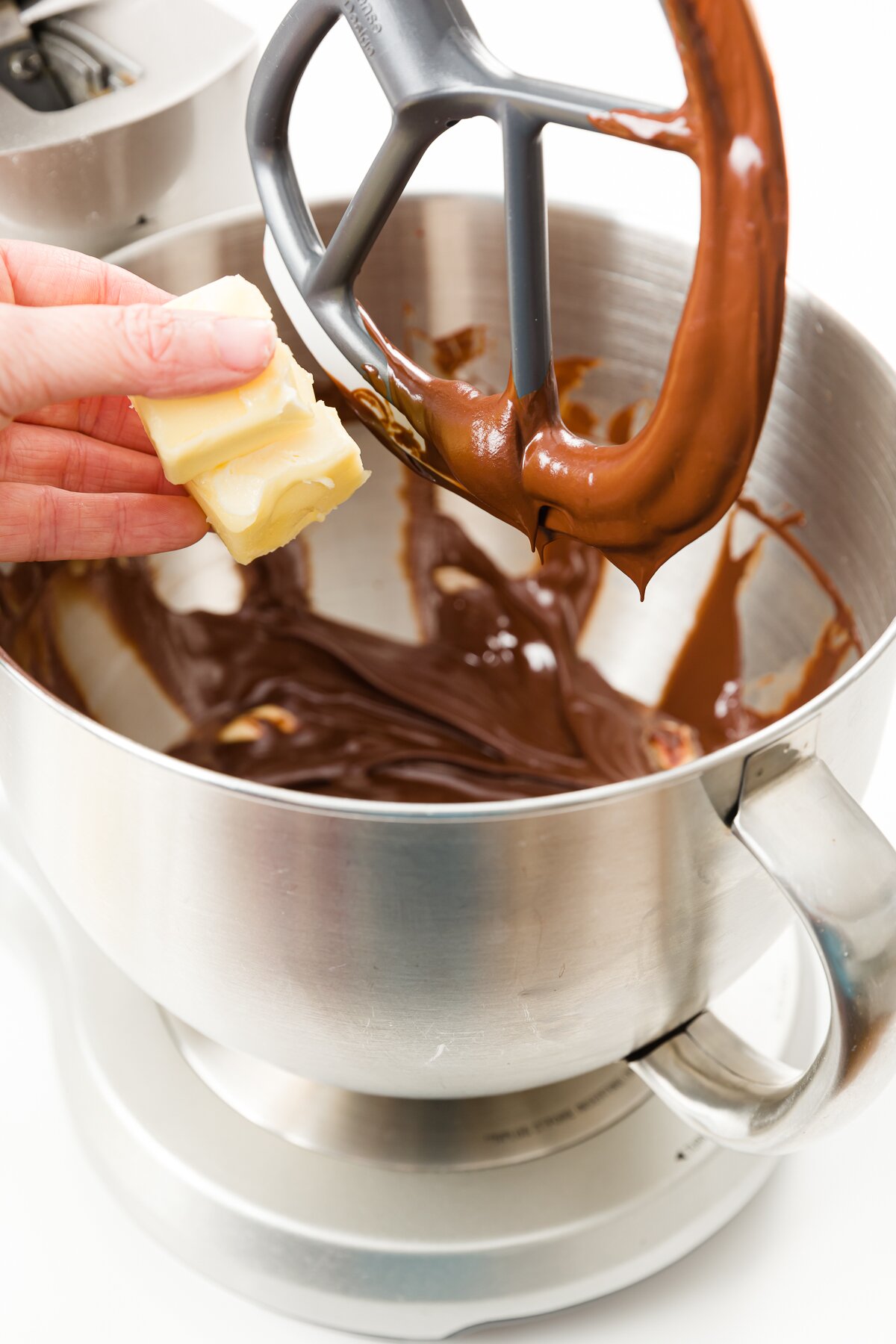 Hand dropping butter pieces into a stand mixer filled with chocolate with paddle lifted