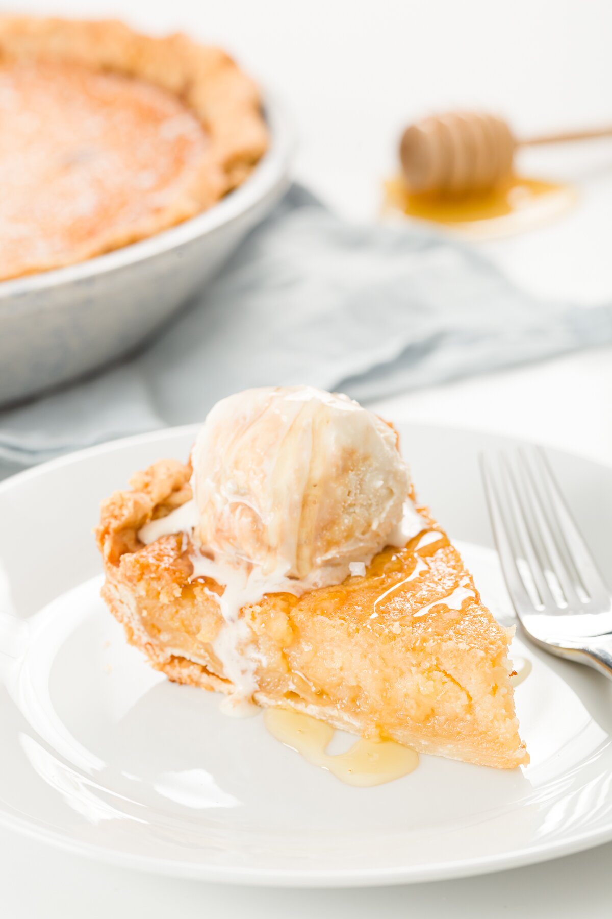Slice of honey pie on a white plate with a scoop of ice cream on top and a pie in the background
