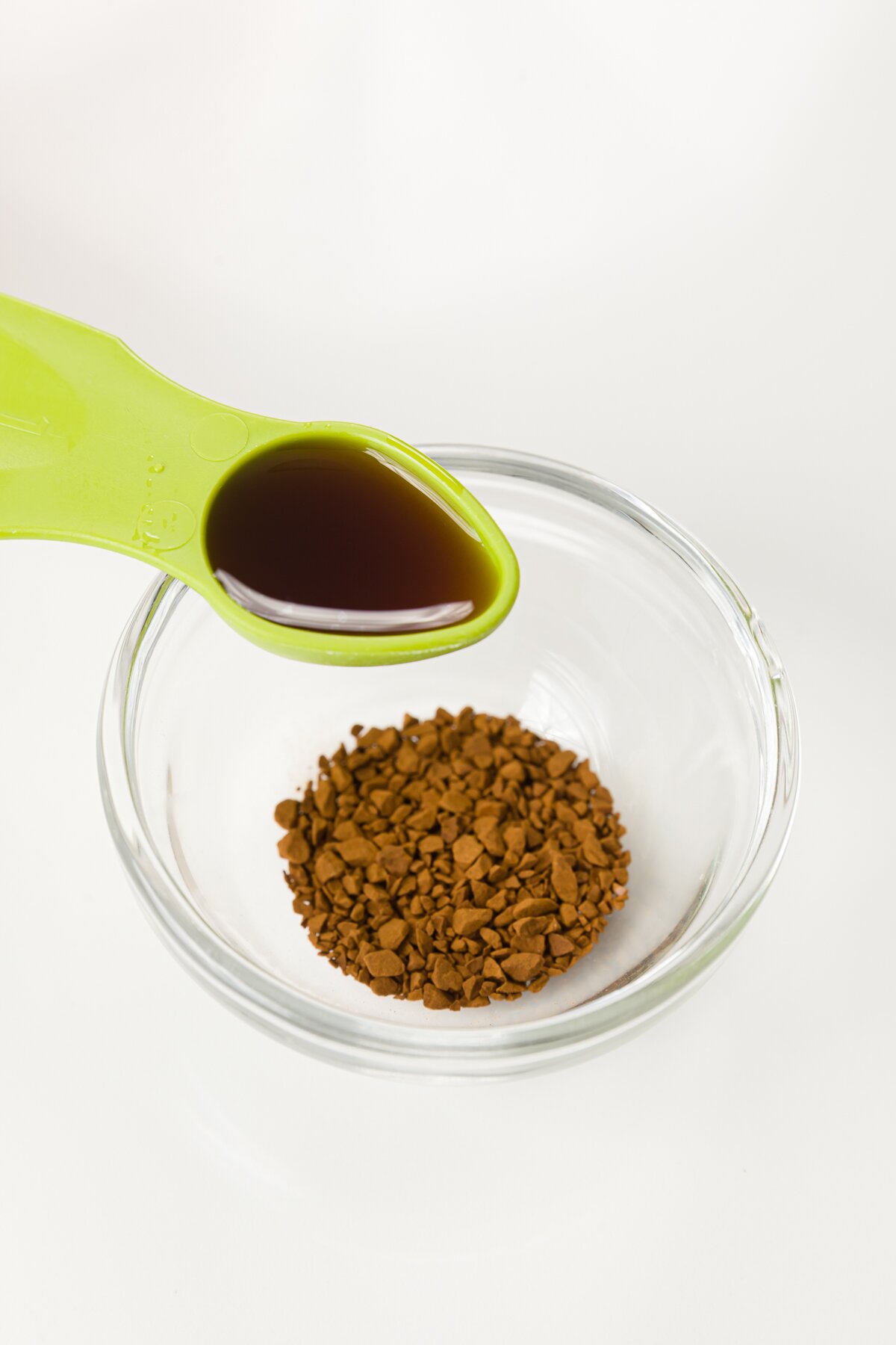 Green tablespoon with vanilla extract held over a glass prep bowl with instant coffee granules in it