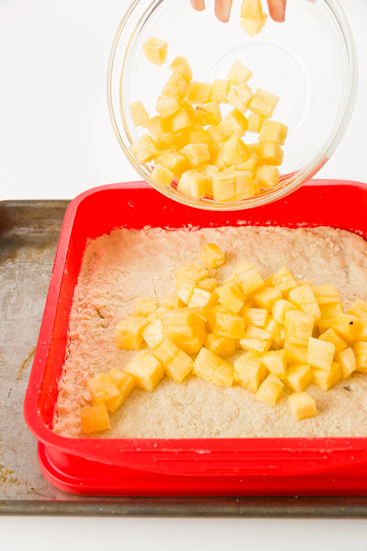 Adding pineapple chunks to the top of a crust in a red baking pan