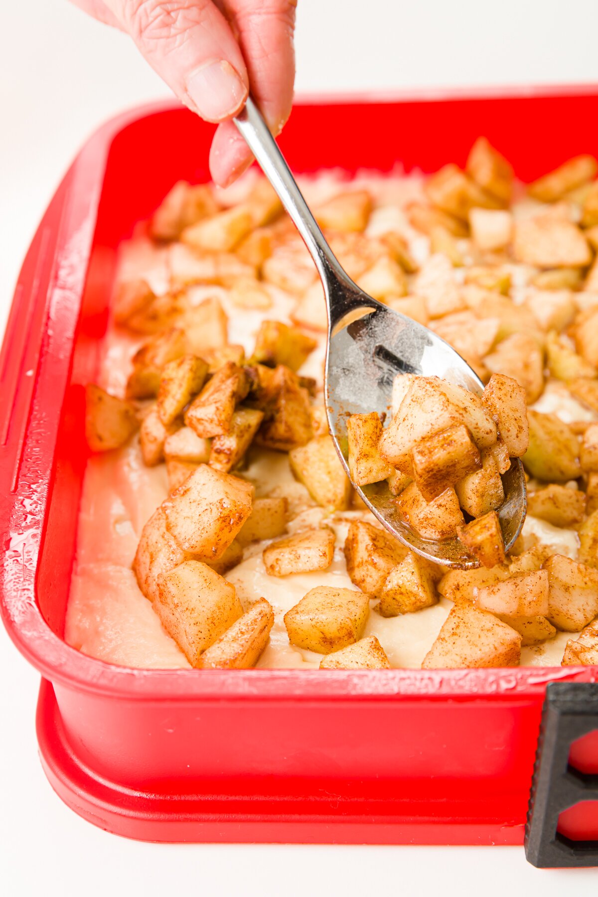 Apple being spread over batter with a spoon in a square pan