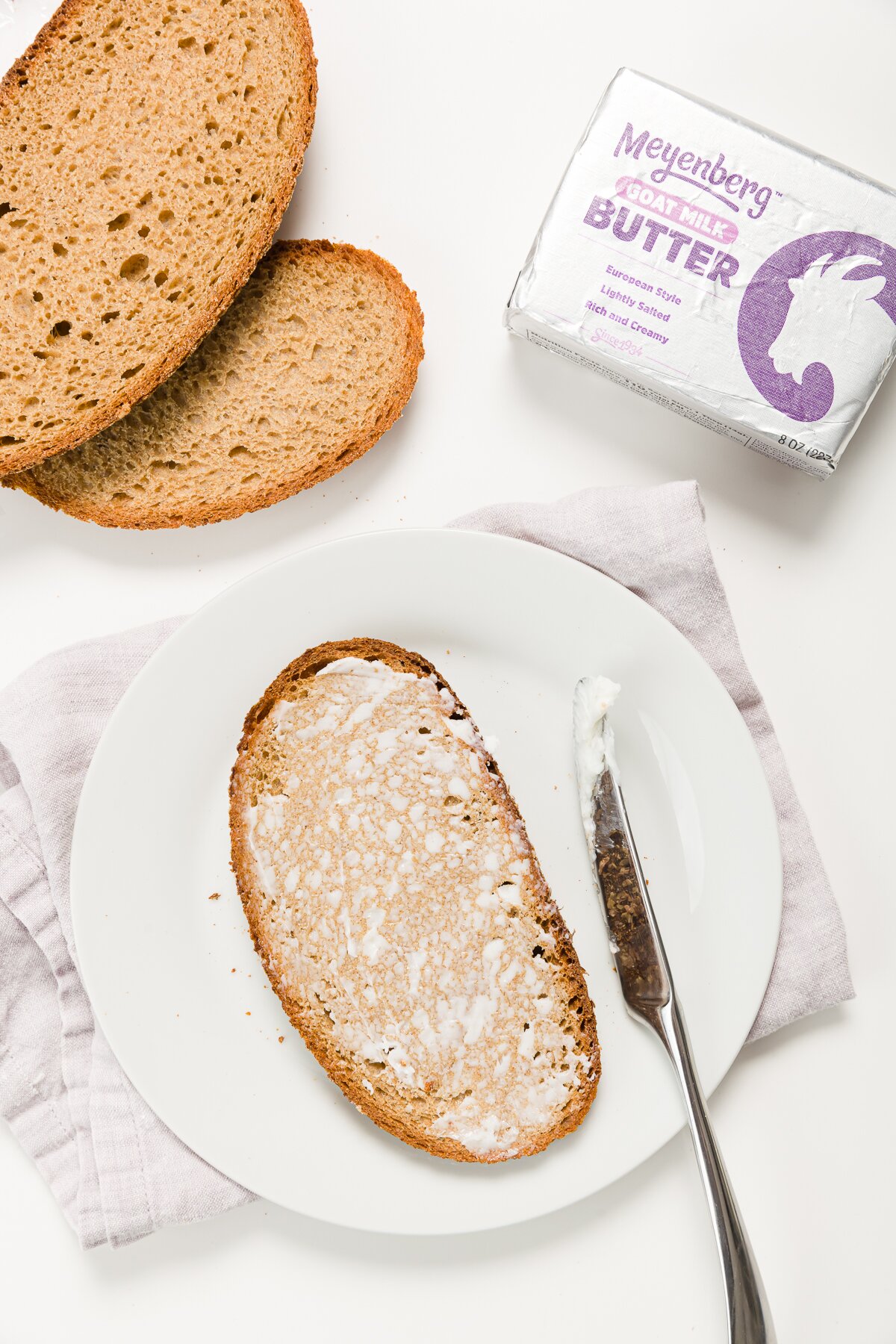 Buttered bread on a plate with a more bread and a package of goat butter in the background