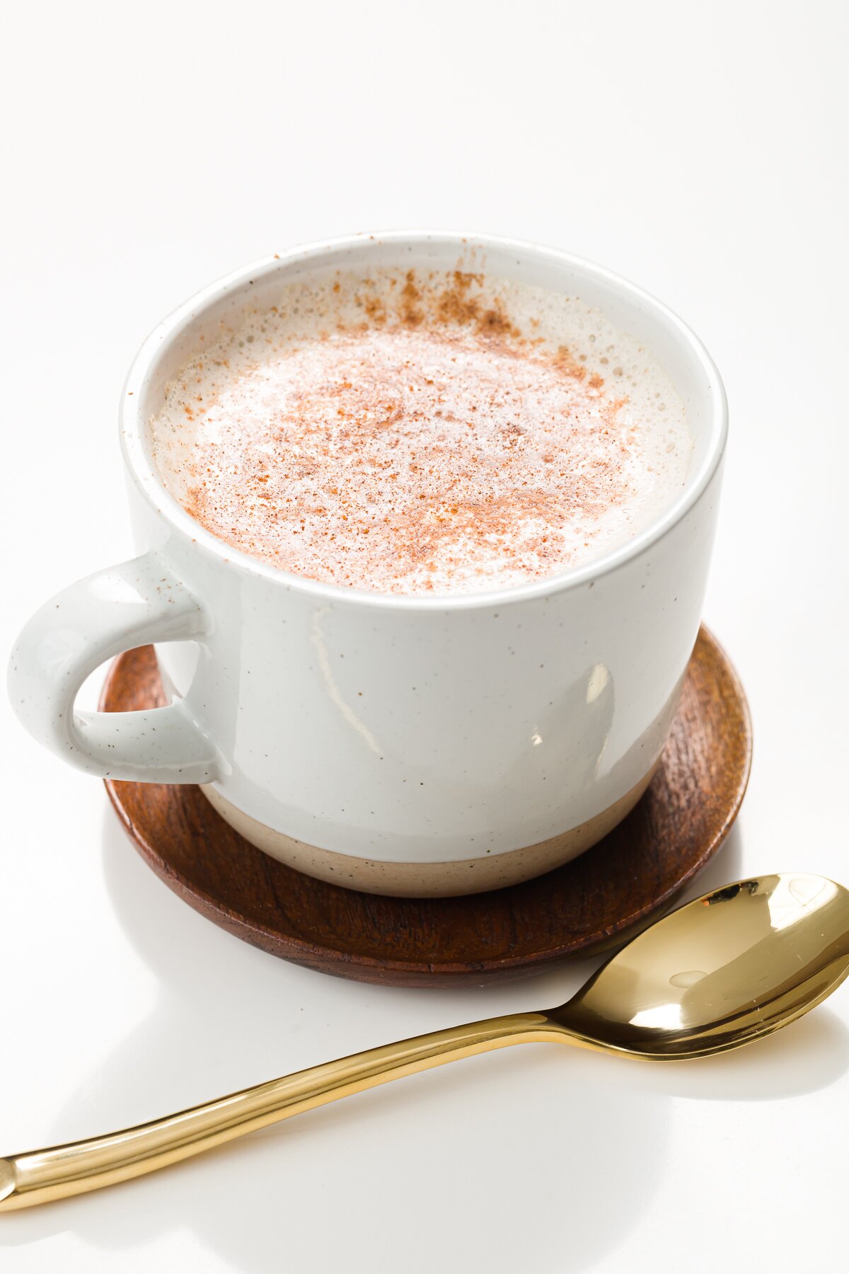 White mug of cinnamon milk on a wood coaster with a gold spoon in front of it